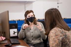 Maggie Keller, with her back to the camera, talks to Carly York, masked, in her office