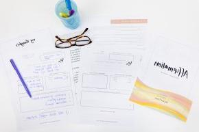 A journal, papers, a pen, cup and glasses sit on a white desk