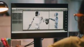 A person sits at a computer that is displaying a robot on the screen