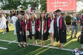 Six students in graduation regalia stand on the field under a sign reading "Congratulations Class of 2024"